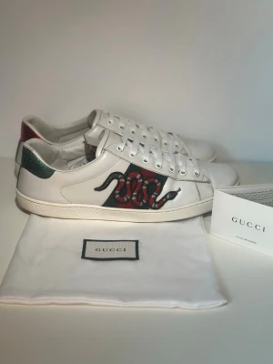 Gucci Ace sneakers med orm - Säljer ett par vita Gucci Ace sneakers i läder med den ikoniska ormbroderingen på sidan och gröna/röda detaljer. Baksidan har metallicgrön och metallicröd hälflik. Klassisk låg modell med vita snören och gummisula. Kommer med dustbag och äkthetskort.