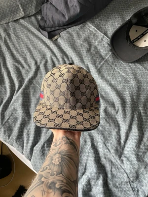 Gucci beige GG keps med röd-blå detalj - Säljer en beige keps från Gucci med klassiskt GG-monogram över hela kepsen och röd-blå rand på sidan. Justerbar rem bak och böjd skärm. Materialet är canvas med insida i mörkt tyg. Perfekt för dig som vill ha en lyxig och trendig accessoar.