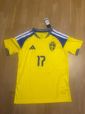 Sveriges vm tröja gyökeres S - Gul fotbollströja från Adidas med blå detaljer, Sveriges landslagsmärke på bröstet och nummer 17 samt namnet Gyökeres på ryggen. Tröjan har korta ärmar, blåa ränder på axlarna och är tillverkad i ett lätt, ventilerande material.