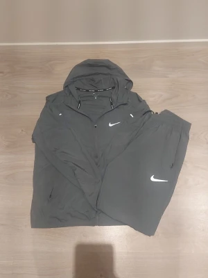 Grå nike miller tracksiut - Säljer ett grått träningsset från Nike med matchande jacka och byxor. Jackan har huva, dragkedja och två fickor, medan byxorna har resår i midjan och sidofickor. Båda plaggen har vita Nike-loggor. Perfekt för träning eller chill.