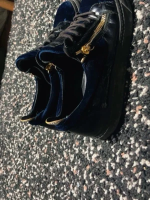 Mörkblå sneakers med gulddragkedja - Säljer ett par mörkblå sneakers i sammet med snygga guldfärgade dragkedjor på sidorna och svarta snören. Skorna har en chunky svart sula och coola detaljer i guld på hälen. Perfekta för dig som vill sticka ut med din stil.