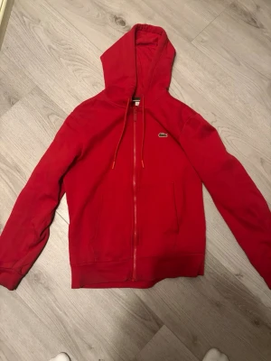 Röd hoodie från Lacoste med dragkedja - Snygg röd hoodie från Lacoste med klassisk passform. Hoodien har dragkedja framtill, två fickor och justerbar huva med snören. Ikoniska Lacoste-loggan på bröstet. Perfekt för en avslappnad och sportig stil.