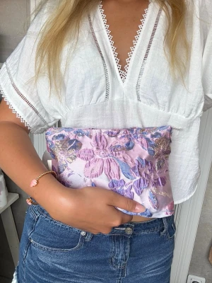 Lila blommig clutch med brodyr - Säljer en liten clutch i lila satin med broderade blommor i rosa, blått och guld. Väskan har dragkedja upptill och en mjuk, rektangulär form. Perfekt för att bära småsaker.