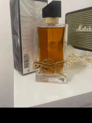 Yves Saint Laurent Libre Eau de Parfum - Säljer en elegant parfym från Yves Saint Laurent, Libre Eau de Parfum. Flaskan är rektangulär i klart glas med guldiga detaljer och en svart asymmetrisk kork. Doftvätskan är bärnstensfärgad och flaskan pryds av YSL:s ikoniska logga i metall.