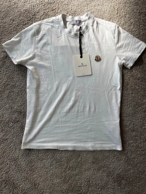Vit Moncler t-shirt med logga - Snygg vit t-shirt från Moncler med klassisk rund hals och korta ärmar. T-shirten har Monclers ikoniska logga broderad på bröstet och är tillverkad i mjuk bomull för en skön känsla. Perfekt basplagg för en clean och stilren look. Ej äkta!!
