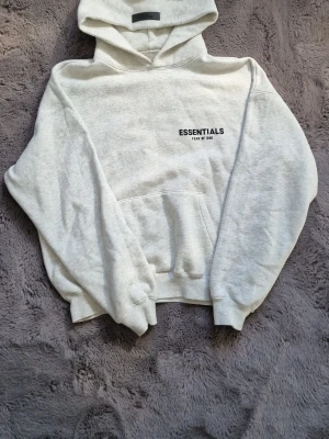 Grå Essentials Fear of God hoodie - Snygg ljusgrå hoodie från Essentials Fear of God med svart logga tryckt på bröstet och ryggen. Hoodien har en stor huva, magficka och ribbade muddar vid ärmar och midja. Tillverkad i mjukt bomullsmaterial för maximal komfort.