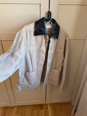 Overshirt med svart skinnkrage från Gina Tricot - Stilren overshirt från Ginatricot, Perfect Jeans  i ljusbeige denim med två stora framfickor och en unik svart krage i skinnimitation. Rak passform och tryckknappar framtill ger en modern look. Perfekt för lager på lager och streetwear-vibe.