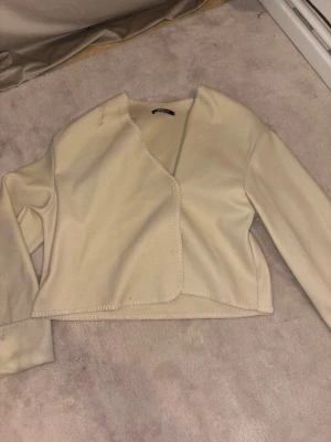Beige croppad kofta med råa kanter - Trendig beige croppad kofta med råa kanter och lång ärm. Koftan har en öppen design utan knappar och är gjord i ett mjukt, fleeceliknande material. Perfekt att slänga över en topp för en avslappnad look.