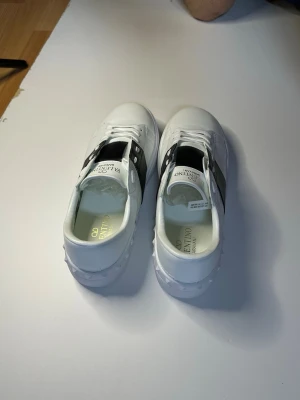 Vita Valentino Garavani sneakers - Snygga vita sneakers från Valentino Garavani med svart band över vristen och vita snören. Skorna har en stilren design med detaljer i guld på insidan och klassisk låg profil. Tillverkade i slätt läder för en clean och exklusiv look.