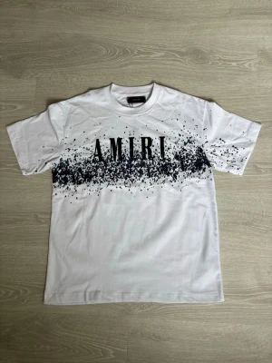 Vit t-shirt med svart print från Amiri - Säljer en vit t-shirt från Amiri med svart stänkmönster och stor logga över bröstet. T-shirten har klassisk passform, rund halsringning och är kortärmad. Materialet är mjuk bomull som känns skön mot huden. Den är stor i storlek 
