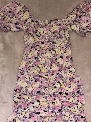 Blommig kortklänning med puffärm - Söt kortklänning med blommigt mönster i lila, rosa, gul och vit. Klänningen har puffiga kortärmar och rynkade detaljer längs hela plagget. Perfekt för sommarens alla tillfällen och ger en romantisk vibe.