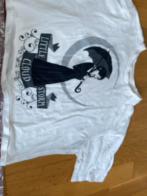Vit Wednesday Addams t-shirt - Vit t-shirt med tryck av Wednesday Addams som håller ett paraply och texten 'Little Storm Cloud' på framsidan. På baksidan står det 'WEDNESDAY' i svart text. T-shirten har rund hals och korta ärmar, perfekt för dig som gillar gotisk stil.