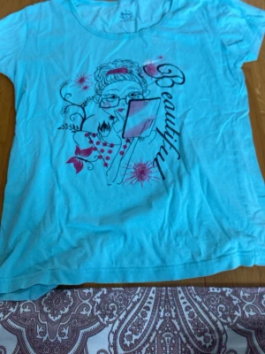 Turkos t-shirt med tryck - Turkos t-shirt med ett coolt tryck av en tjej med glasögon och texten 'Beautiful' i svart. T-shirten har rund halsringning och korta ärmar. Materialet känns mjukt och skönt, troligen bomull. Perfekt för dig som gillar färg och grafiska motiv.