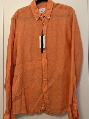 Orange linneskjorta - Bläck - Snygg orange skjorta i linne från Bläck med klassisk krage och vita knappar. Skjortan har lång ärm och en avslappnad passform, perfekt för varmare dagar. Enkel och stilren design som funkar till många olika looks.