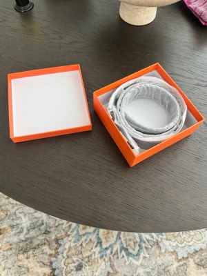 Svart skärp med silverdetaljer - Snyggt svart bälte i skinn med silverfärgat spänne. Bältet har en stilren design med vita sömmar längs kanterna och levereras i en orange presentask. Perfekt accessoar för att lyfta din outfit.