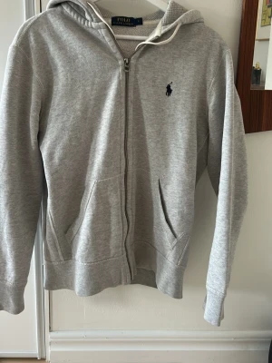 Grå hoodie från Polo Ralph Lauren - Snygg grå zip hoodie från Polo Ralph Lauren. Passar dig som är runt 170 cm. Välanvänd men fortfarande i gott skick.