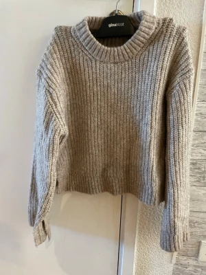 Beige stickad tröja med slit i ärmarna Gina Tricot  - Mysig beige stickad tröja från Gina Tricot med grov ribbstickning. Tröjan har en hög, rund halsringning och långa ärmar med breda muddar. Perfekt för lager på lager och enkel att matcha till olika outfits.