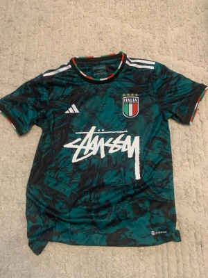 Italien fotbollströja Stüssy x Adidas - Unik grön och svart fotbollströja från Adidas i samarbete med Stüssy och Italien. Tröjan har ett coolt mönster, Italiens landslagsemblem med fyra stjärnor, vita Adidas-ränder på axlarna och Stüssy-logga i vitt framtill. Ribbade detaljer i Italiens färger vid hals och ärmslut.