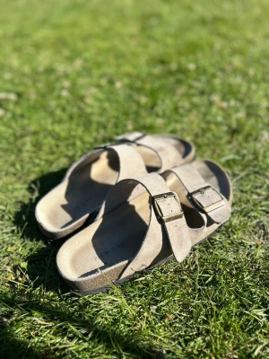 Beige sandaler från Pier one - Snygga beige sandaler från pier one med två breda remmar i mocka och justerbara metallspännen. Klassisk fotbädd som ger skön komfort och en stilren look. Perfekta för varma dagar och enkel att matcha med olika outfits.