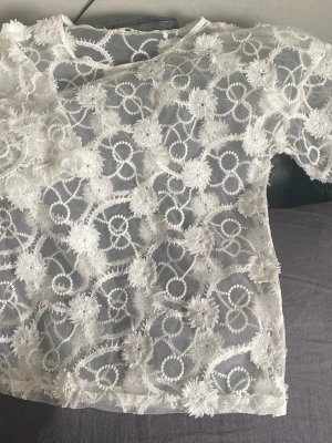 Vit blommig spetsblus - Säljer en superfin vit blus i transparent mesh med broderade blommor och swirl-detaljer. Blusen har korta ärmar och är perfekt för dig som gillar romantisk och unik stil. Materialet känns lätt och luftigt, och spetsen ger en lyxig känsla.