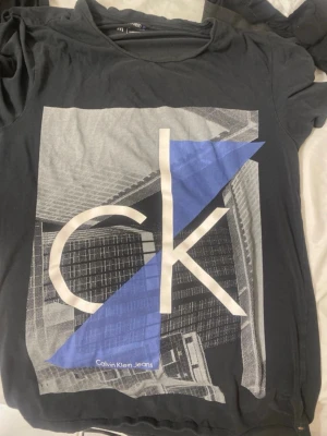 Svart Calvin Klein Jeans t-shirt - Snygg svart t-shirt från Calvin Klein Jeans med stort grafiskt tryck i vitt, grått och blått framtill. Klassisk rund hals och korta ärmar. Tillverkad i mjuk bomull med stretch för extra komfort. Perfekt för en trendig och avslappnad look.