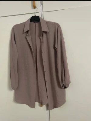 Beige oversized skjorta med knappar - Lång, oversized beige skjorta med knappar framtill och långa, lösa ärmar med lätt puff vid mudden. Skjortan har klassisk krage och är tillverkad i ett mjukt, strukturerat tyg som ger en avslappnad look.
