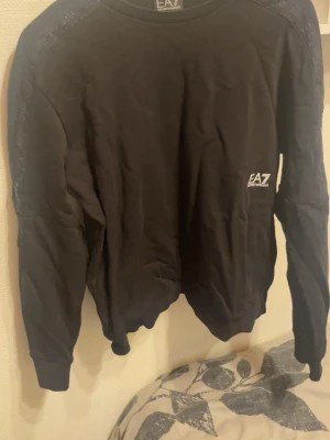 Svart sweatshirt från EA7 Armani - Svart sweatshirt från EA7 Emporio Armani med rund halsringning och diskret logga på bröstet. Tröjan har ribbade muddar vid ärmslut och nederkant samt detaljer längs axlarna. Perfekt för en stilren och sportig look.
