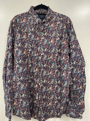 Blommig skjorta från Grand Frank - Blommig skjorta från Grand Frank med färgstarkt mönster i blått, gult, grönt och rött på mörk botten. Skjortan har klassisk krage, knappar framtill och långa ärmar. Perfekt för dig som vill sticka ut med en unik stil.