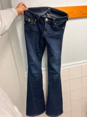 Blå bootcut jeans från True Religion - Säljer ett par mörkblå bootcut jeans från True Religion med kontrastsömmar och snygga fickdetaljer bak. Klassisk femficksmodell med silverfärgade knappar och logopatch bak i midjan. Tillverkade i stretchig denim för skön passform. Slitet baksida längst ner