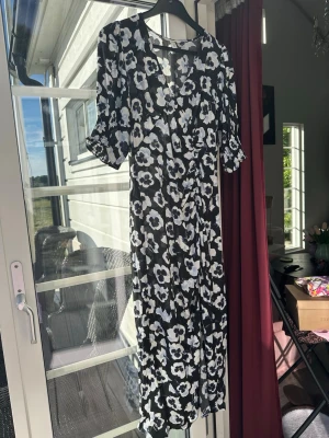 Svart blommig långklänning H&M - Svart långklänning med vita och ljusblå blommor, v-ringad och med korta puffärmar. Klänningen har en lätt rynkad detalj i midjan och är tillverkad i ett tunt, luftigt material som passar perfekt för varmare dagar. Slits nertill på ena sidan. Storlek M