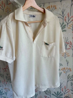 Beige pikétröja från Lacoste - Klassisk beige pikétröja från Lacoste med kort ärm, krage och knappslå. Tröjan har en bröstficka med den ikoniska krokodilloggan och är tillverkad i bomull. Perfekt för en avslappnad och stilren look M/L.