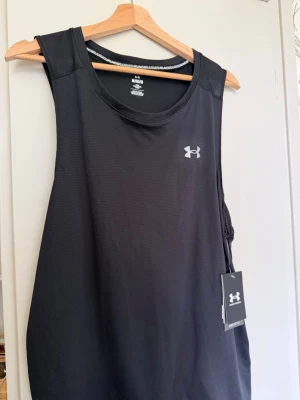 Svart träningslinne från Under Armour - Snyggt svart träningslinne från Under Armour med rund halsringning och diskret logga på bröstet. Tillverkat i ett lätt och ventilerande funktionsmaterial som passar perfekt för gymmet eller löprundan. Klassisk passform och sportig look.