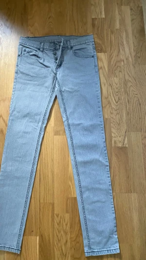 Grå Cheap Monday "Tight" – Low/Mid waist (Y2K/Indie) - Modell: Skinny jeans med normalhög midja (mid waist) och knappgylf.Skick: Mycket fint skick, använda endast 2 gånger. Notera att märkeslappen baktill har lossnat lite i kanten.Material: 98% Bomull, 2% Spandex (ger stretch).Storlek & Mått (tagna platt)Märkt storlek: W28 / L32 (motsvarar Small / 36).Midjemått: ca 38 cm (76 cm omkrets).Innerbenslängd: ca 79 cm.Grenhöjd: ca 20 cm.