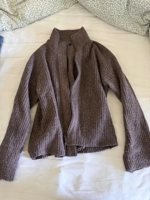 Brun ribbad ziptröja med hög krage - Mysig brun ribbad tröja med dragkedja framtill och hög krage. Tröjan har långa ärmar och är tillverkad i ett mjukt stickat material som ger en avslappnad look. Perfekt för lager på lager och chill dagar.