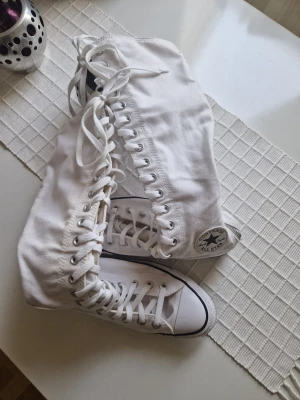 Vita höga Converse All Star sneakers - Säljer ett par vita Converse All Star sneakers med högt skaft och klassisk svart logga på sidan. Skorna har vita snören, tåhätta i gummi och sula med svart rand. Tillverkade i canvas som ger en clean och tidlös look.