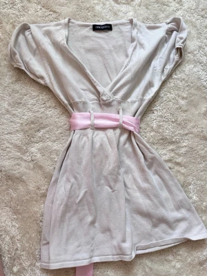 Ljusgrå babydoll med rosa sidenband - Supersöt ljusgrå babydoll från New Fashion med korta puffärmar och djup v-ringning. Midjan markeras av ett brett rosa sidenband som knyts i en rosett bak. Perfekt för att ge outfiten en romantisk touch. Mjukt och tunt stickat material.