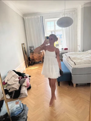 Vit kort sommarklänning med tunna band - Söt vit kort klänning med tunna axelband som går och justera och volangdetaljer. Klänningen har en luftig passform och är perfekt för varma dagar. Tillverkad i ett lätt tyg som ger en avslappnad och fräsch look.