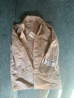 Beige trenchcoat från Acne Studios - Stilren beige trenchcoat från Acne Studios med klassisk krage och dubbelknäppning. Jackan har en stor vit patch med logga på ärmen och detaljer i form av ljusa knappar. Materialet är tunt och lätt, perfekt för lager på lager och en modern look.