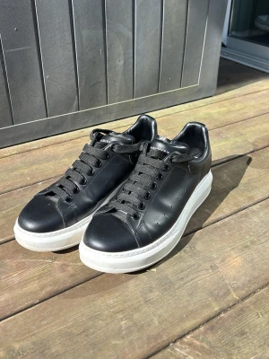 Svarta Alexander McQueen sneakers - Stilrena svarta sneakers från Alexander McQueen med chunky vit sula och svarta skosnören. Skorna är i slätt läder och har diskret perforering på sidorna samt logga på hälen. Perfekta för dig som gillar en clean och modern look.