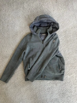 Grå stickad cardiganjacka Moncler - Snygg grå stickad cardiganjacka från Moncler med dragkedja, två fickor med dragkedja och en vadderad huva. Jackan har Moncler-logga på bröstet och ribbade muddar. Perfekt för lager på lager och streetwear-vibe.