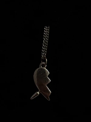 Halsband med trasigt hjärta-hänge - Säljer ett halsband med ett silverfärgat hänge i form av ett trasigt hjärta. Hänget har en kantig, sicksackad spricka och sitter på en tunn kedja. Köpt här på plik för 200