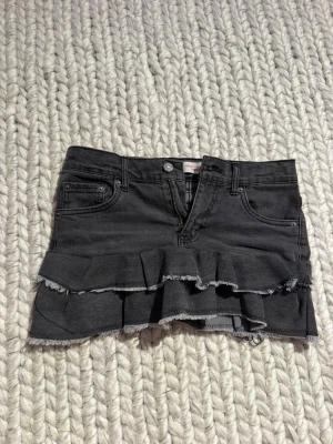 Svart volangkjol i denim från Levis - Cool svart/grå jeanskjol från Levis med volangdetaljer och råa kanter. Kjolen har klassisk femficksdesign, bälteshällor och dragkedja fram. Perfekt för dig som gillar en edgy och trendig stil.