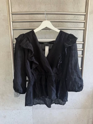 Svart volangblus från Only - Trendig svart blus från Only med v ringning, volangdetaljer längs axlar och bröst samt markerad midja. Blusen har långärmad design med lätt puffade ärmar och är tillverkad i ett tunt, strukturerat tyg som ger en luftig känsla.