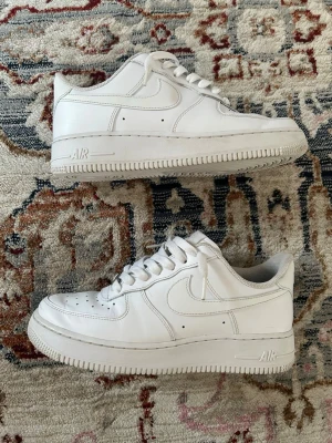 Nike Air Force 1 vita sneakers - Klassiska vita Nike Air Force 1 sneakers i lågt utförande. Skorna har en stilren design med perforerad tå, vita skosnören och den ikoniska swoosh-loggan på sidan. Tillverkade i läder med en rejäl sula och AIR-dämpning för extra komfort.