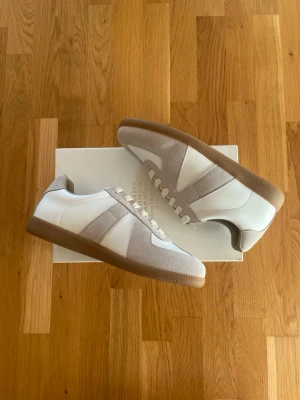 Vita Maison Margiela GATs - Säljer ett par ikoniska Maison Margiela Sneakers i den klassiska färgställningen vitt läder och beige mocka med brun gummisula. Originalkartong medföljer självklart (syns på bilden) 📦. Skriv vid frågor eller om du vill ha fler bilder! Svarar snabbt. 🤝