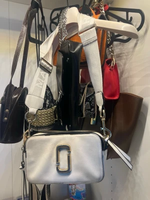 Vit handväska från Marc Jacobs - Säljer en snygg vit handväska från Marc Jacobs med bred axelrem och silverfärgade detaljer. Väskan har en rektangulär form och ett stort metallogo framtill. Perfekt för dig som gillar trendiga accessoarer och vill sticka ut.