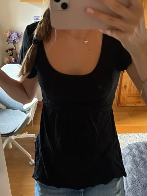 Svart topp med knyt i midjan - Snygg svart topp med korta ärmar och rund, djup urringning. Toppen har ett knytband i midjan som ger en fin siluett och är tillverkad i ett mjukt bomullsliknande material. Passar perfekt till jeans eller kjol för en enkel men stilren look.