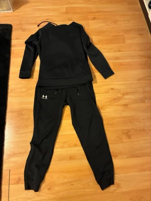 Svart träningsset från Under Armour - Snyggt svart träningsset från Under Armour med matchande hoodie och byxor. Hoodien har en klassisk känguruficka och byxorna har dragsko i midjan samt ribbade benslut. Perfekt för gymmet eller chill. Materialet är mjukt och stretchigt för maximal komfort.