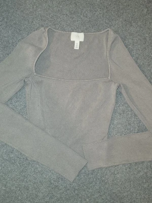 Grå ribbad långärmad topp H&M - Snygg grå ribbad topp från H&M med lång ärm och fyrkantig ringning. Toppen har en slim passform och är tillverkad i mjuk bomullsmix som känns skön mot huden. Perfekt att styla med jeans eller kjol för en clean look.