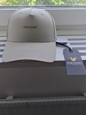 Beige keps från Lyle & Scott - Snygg beige keps från Lyle & Scott med klassisk böjd skärm och broderad logga framtill. Justerbar baktill för perfekt passform. Perfekt accessoar för en clean och stilren look.
